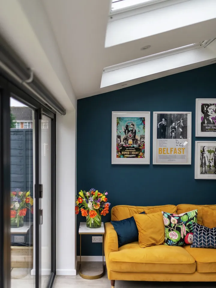 Koyu mavi duvarlı, desenli yastıklarla süslenmiş hardal sarısı bir kanepe, göz alıcı sanat eserleri ve doğal ışığın aydınlatmasına izin veren pencereler ile bir oturma odası.