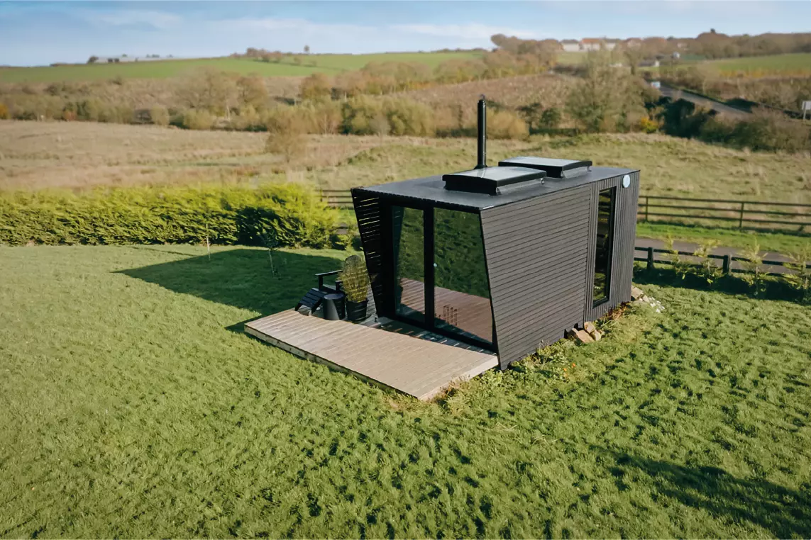 Petite cabane moderne noire avec de grandes fenêtres et une cheminée, située au milieu d’un champ vert avec des haies et des arbres en arrière-plan sous un ciel clair.