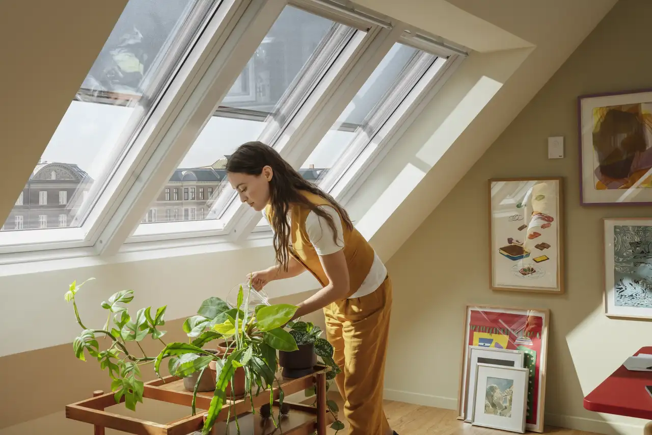 Een vrouw geeft planten water onder dakvensters.