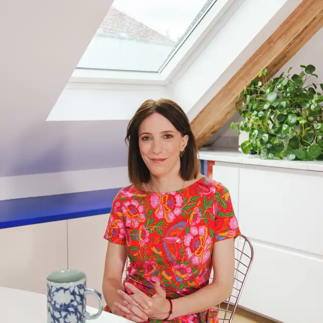 Žena sedící u střešního okna VELUX s šálkem čaje v barevném květinovém oblečení a užívá si přirozeného světla. | VELUX