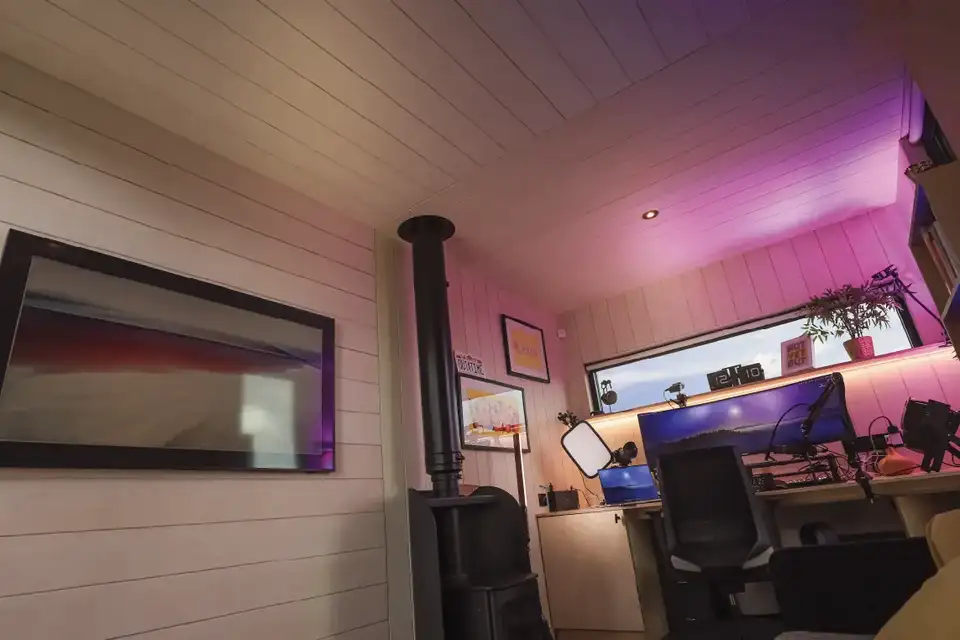 Intérieur avec plafond incliné, suggérant une pièce sous les combles. À gauche, un poêle à bois noir avec un tuyau vertical. Au centre, un grand moniteur sur un bureau avec divers objets. Éclairage ambiant rose et violet, créant une atmosphère cosy.