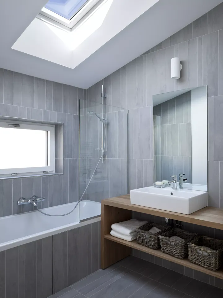Maison Air et Lumière’in modern banyosu, gri fayanslarla döşenmiş ve duş ile küvetin üzerinde bulunan VELUX çatı penceresi sayesinde bol doğal ışık alan bir alan sunuyor | VELUX.