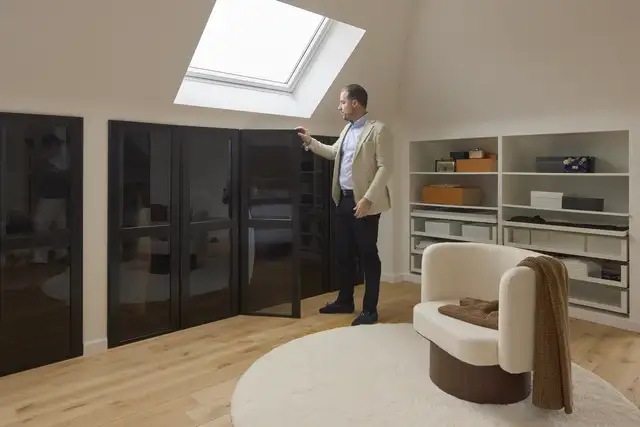 Mężczyzna stojący w nowoczesnej sypialni na poddaszu z dużym oknem dachowym i eleganckimi czarnymi szafami.