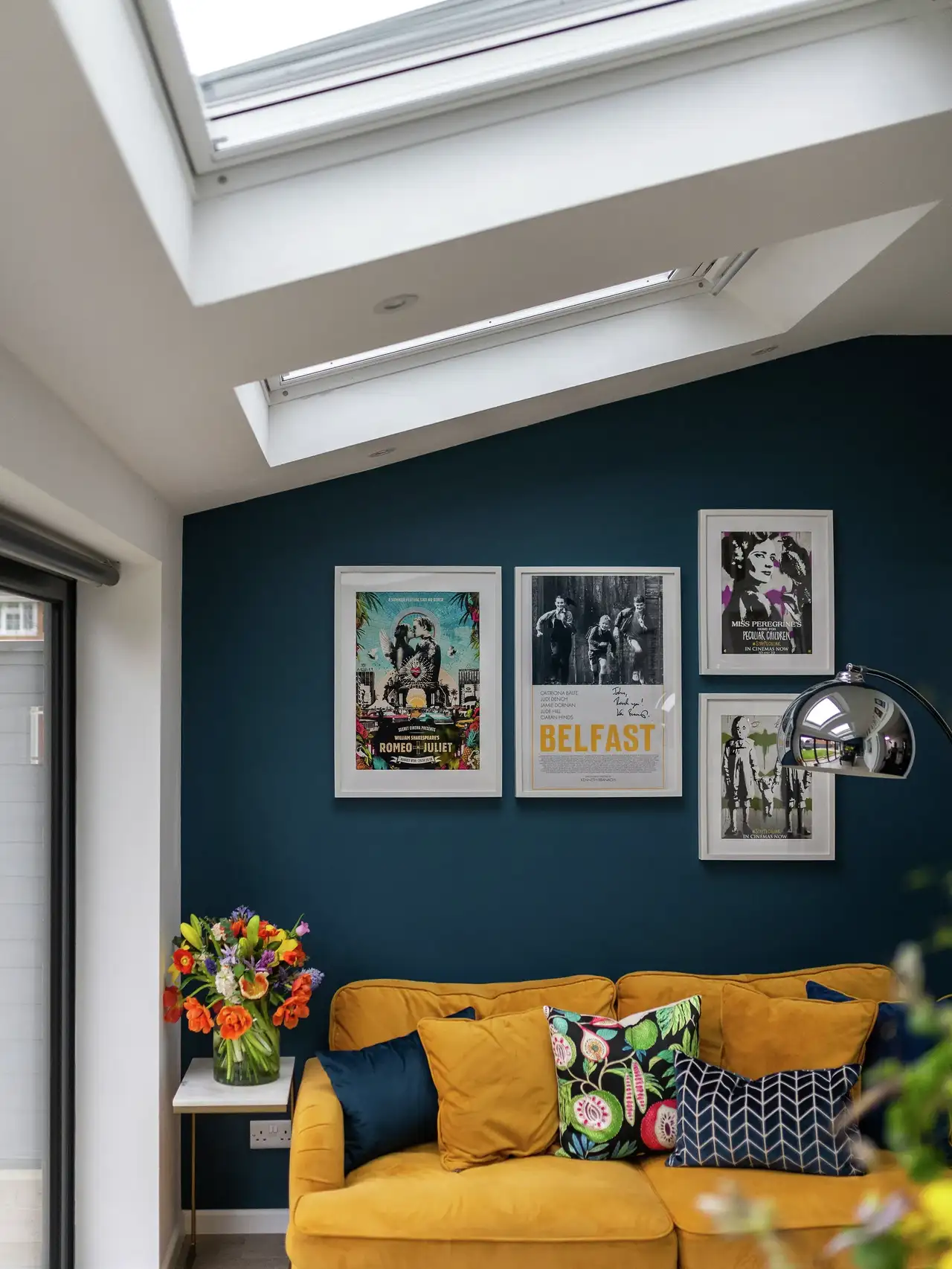 Salon avec mur bleu foncé, canapé jaune moutarde et coussins colorés, éclairé par des fenêtres de toit.