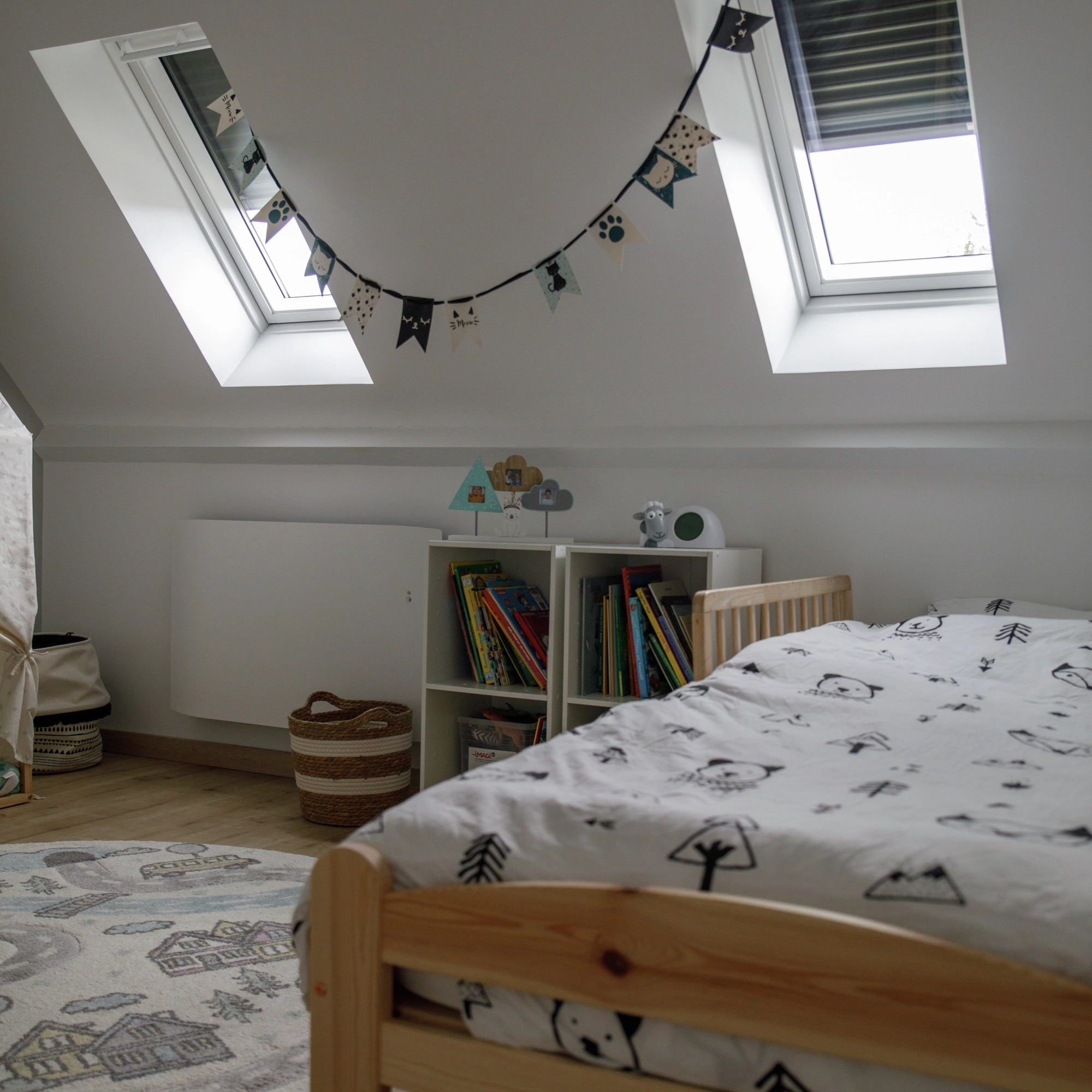 Chambre d’enfant lumineuse avec deux fenêtres de toit inclinées, un lit en bois, une bibliothèque remplie de livres et une guirlande décorative suspendue.