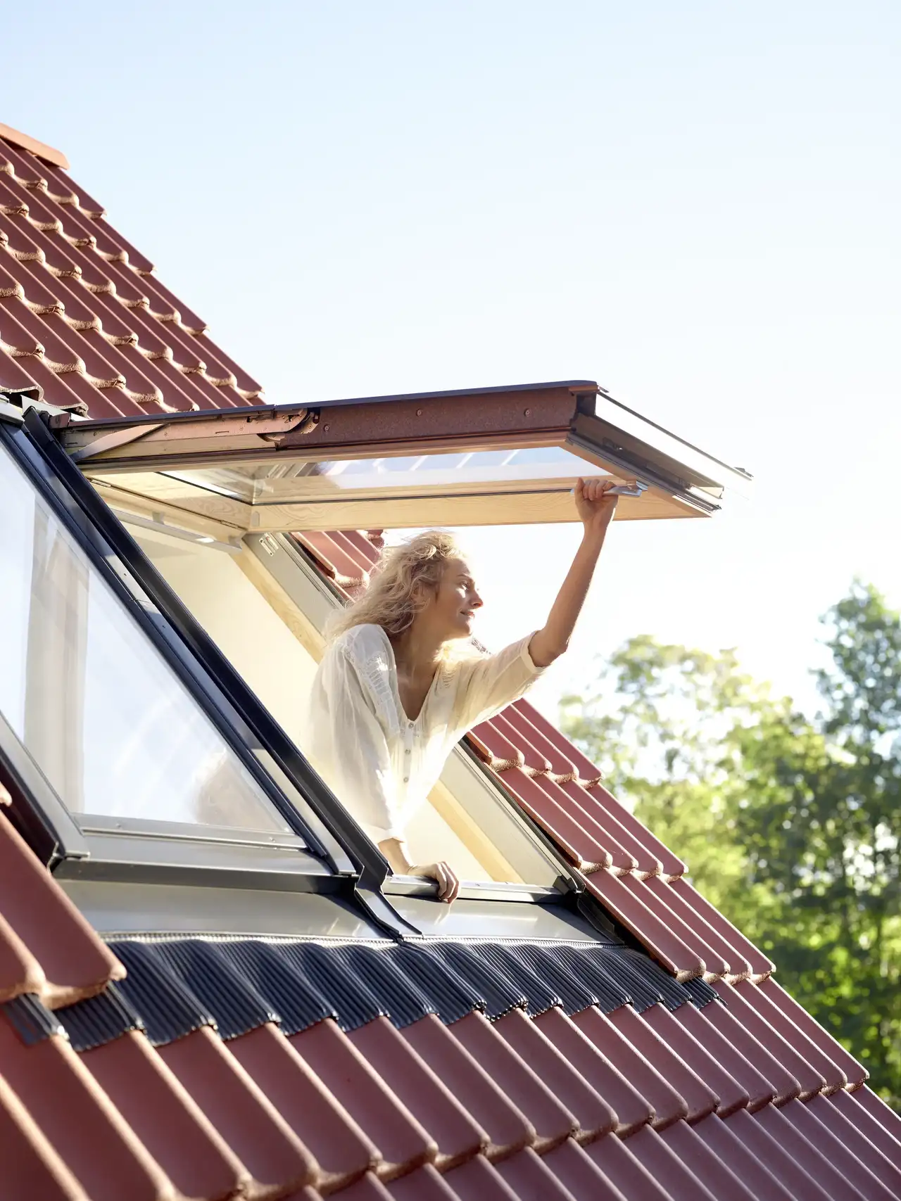 Femme ouvrant une fenêtre de toit VELUX sur un toit en tuiles rouges 