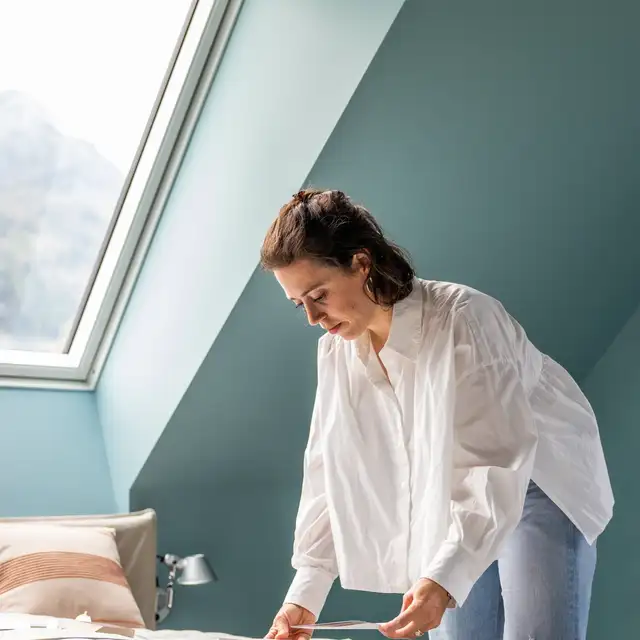 Žena stojící v dobře osvětlené ložnici pod střešními okny VELUX a pečlivě zkoumající vzorky barev na posteli. | VELUX