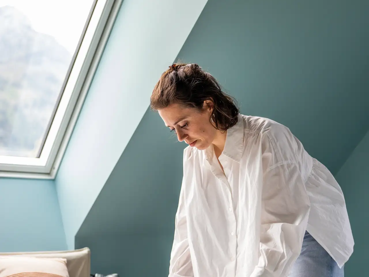 Egy nő dolgokat igazgat az ágyon egy hálószobában, amelyet egy nagy tetőablakon beáradó természetes fény tölt be. | VELUX