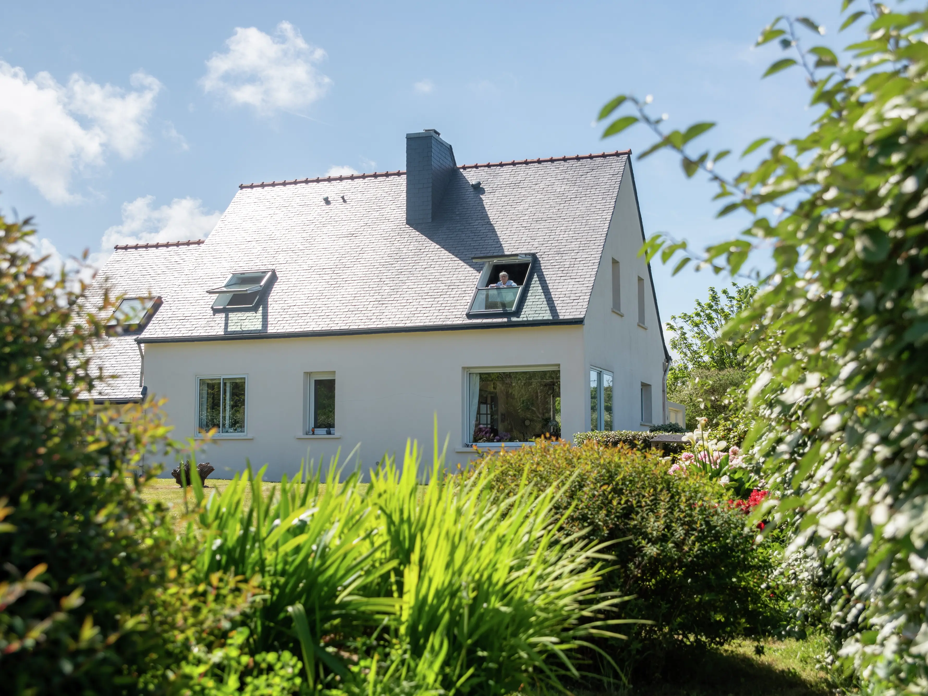 Maison individuelle blanche avec fenêtre de toit VELUX, entourée d'un jardin sous un ciel bleu.
