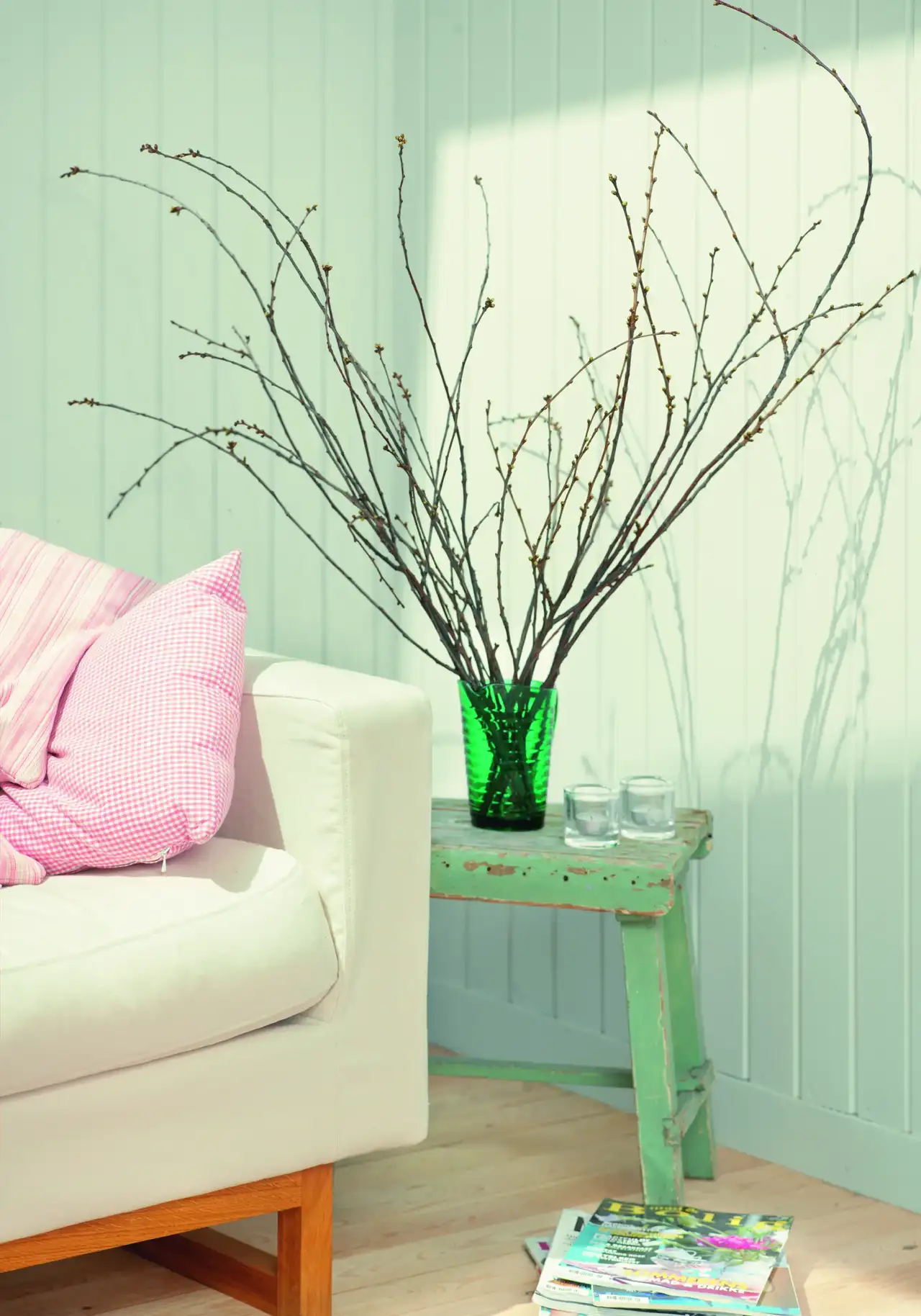 Stillleben im Danish-Pastel-Stil mit grüner Glasvase und Zweigen auf einem Vintage-Hocker, daneben pastellrosa Kissen auf einem hellen Sofa vor weißer Holzvertäfelung | VELUX Magazin