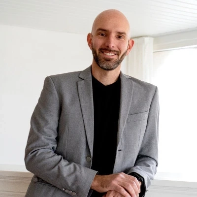 Smiling man in a grey blazer leaning on a white railing in a bright room