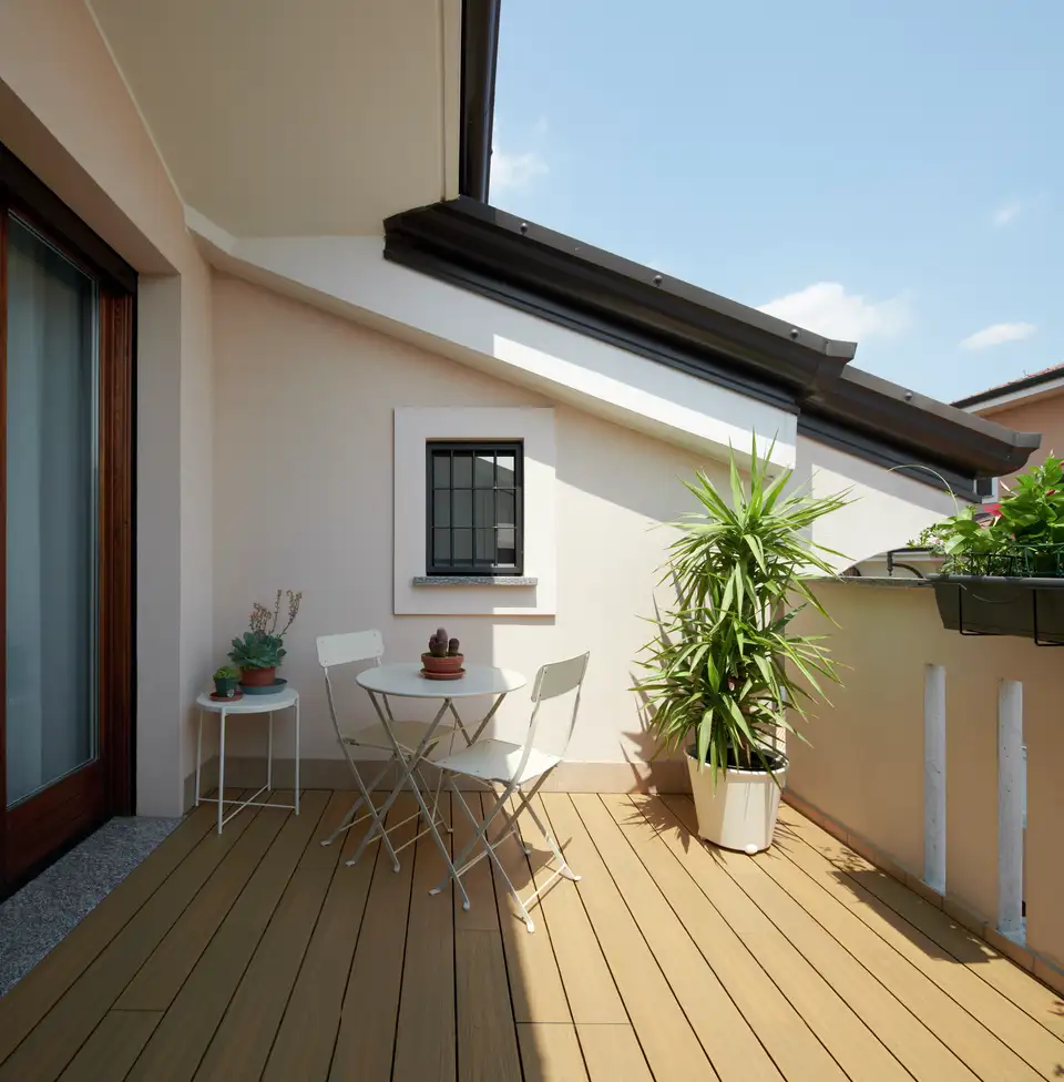 L’image montre un petit balcon confortable avec un plancher en bois. Il y a une table blanche accompagnée de quatre chaises, et diverses plantes sont disposées autour de l’espace. Une fenêtre de toit inclinée au-dessus suggère que ce balcon fait partie d’un grenier ou d’un étage supérieur avec un toit en pente. Cet espace semble conçu pour la détente ou des réunions intimes en plein air.
