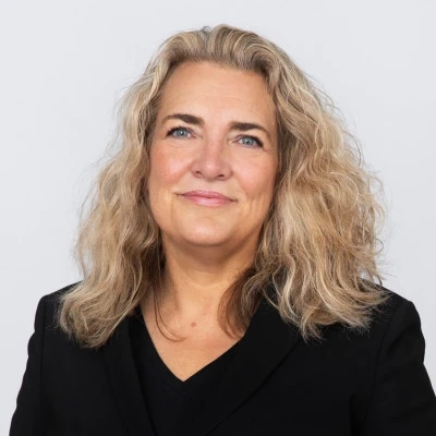 Woman with wavy blonde hair in a black blazer against a plain light background