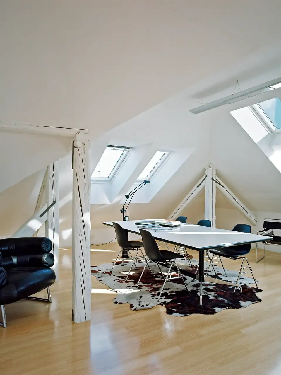 Grenier lumineux aménagé avec table rectangulaire, chaises et fauteuils noirs sur sol clair.