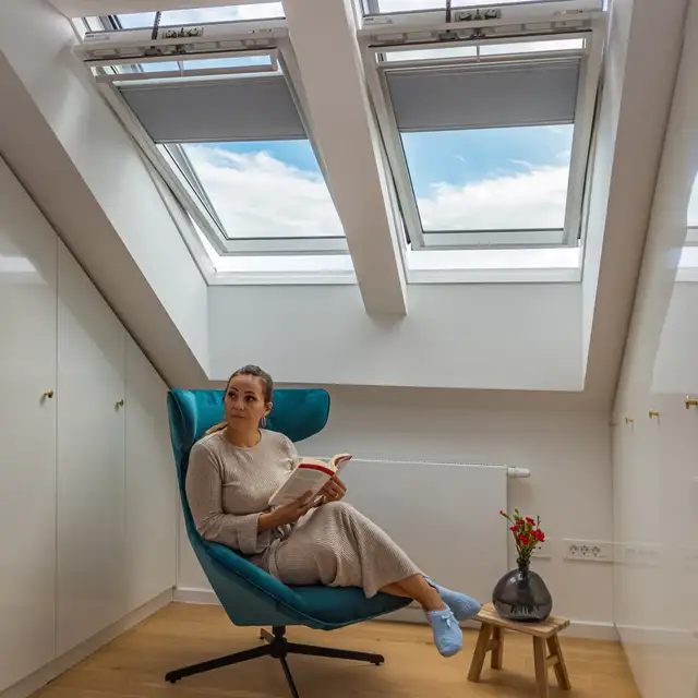 Pohodlný čitateľský kútik so ženou sediacou v kresle pod strešnými oknami a držiacou knihu. | VELUX