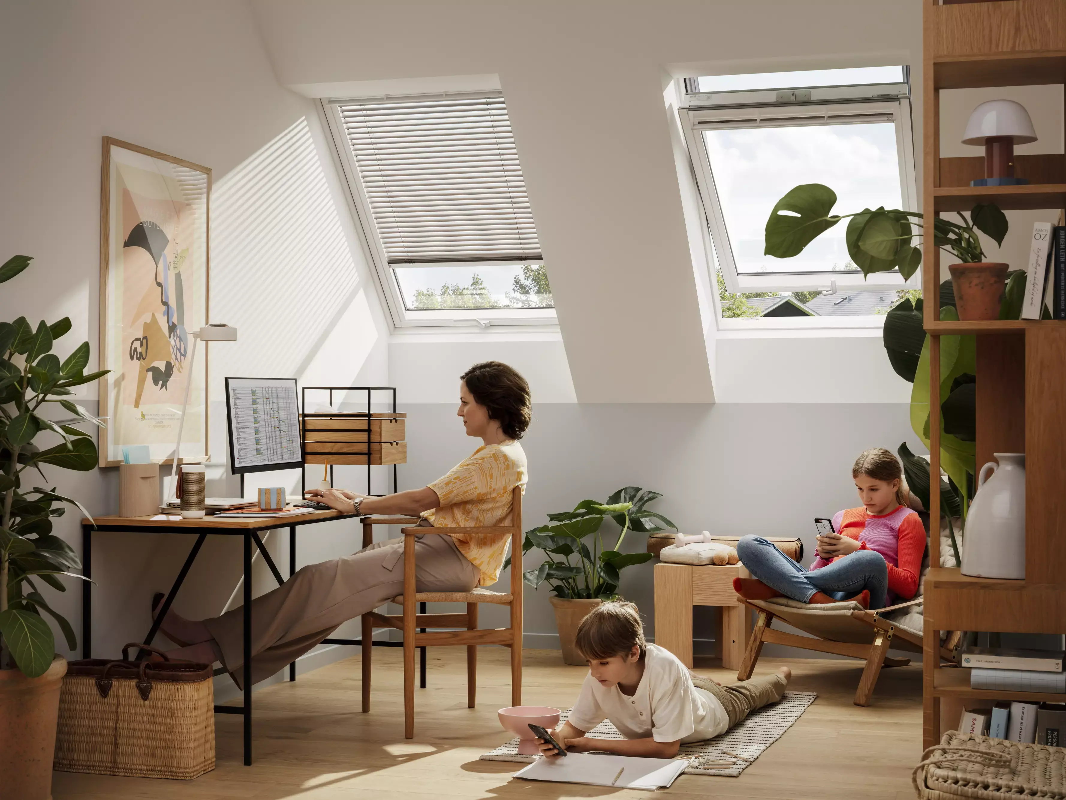 Bureau à domicile sous les combles avec des fenêtres VELUX, une personne au bureau, une autre sur une chaise avec un téléphone, un enfant qui dessine.