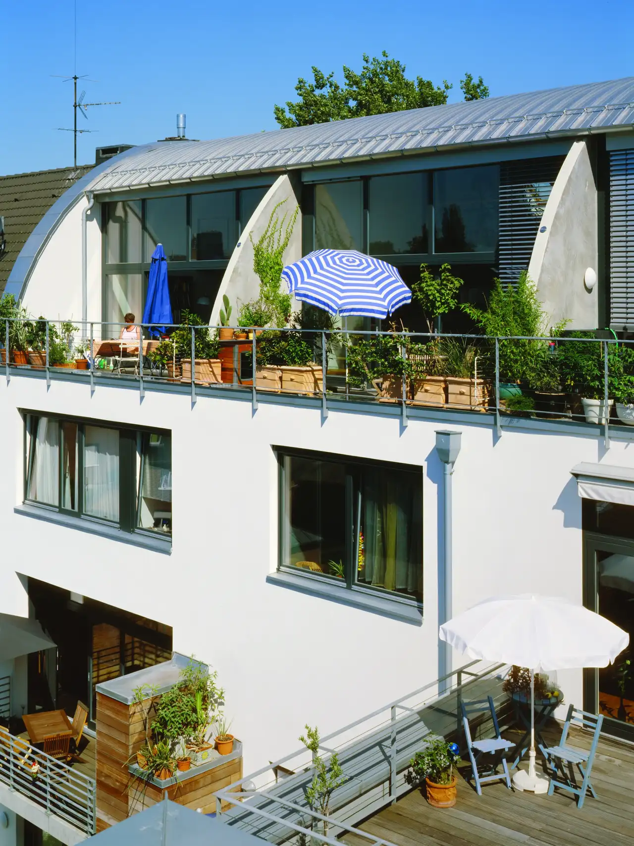 Un bâtiment moderne doté d'un toit arqué unique, avec un jardin en terrasse sur le toit. La terrasse est agrémentée de plantes, de mobilier d'extérieur et de parasols, et constitue un exemple d'utilisation d'un espace vert urbain.