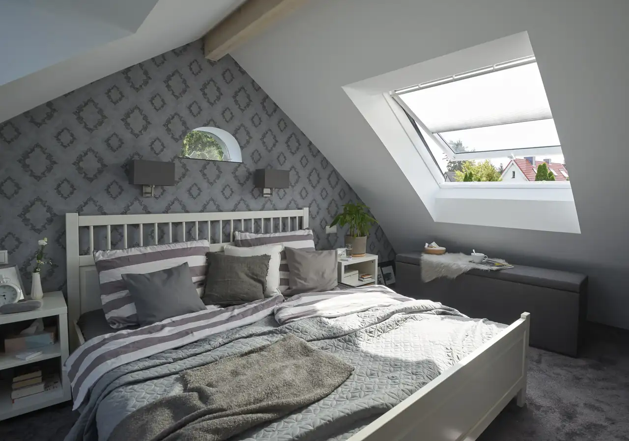 Chambre mansardée avec mur en pente tapissé de papier peint graphique gris et grande fenêtre de toit.
