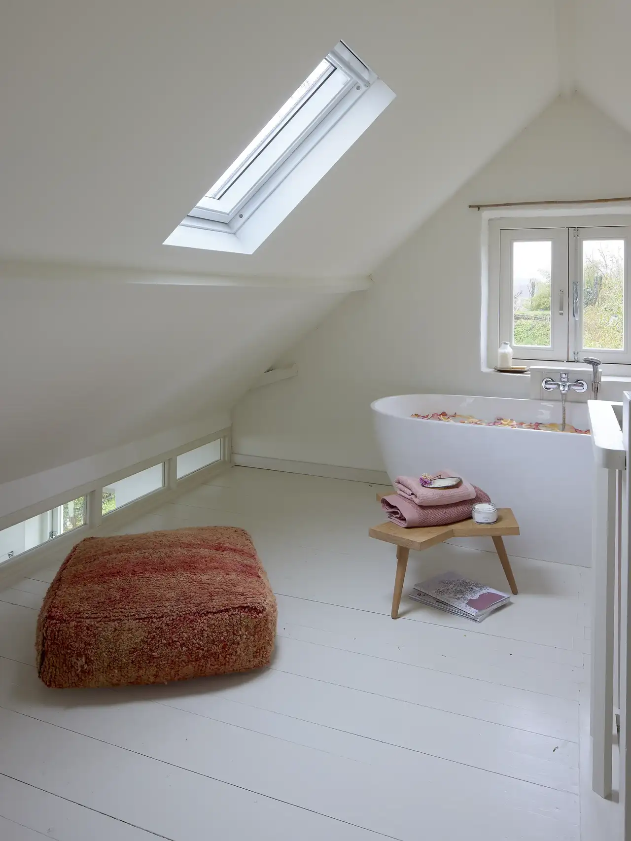 Vasca da bagno freestanding riempita di petali di rosa galleggianti accanto a uno sgabello con asciugamani e una crema | Rivista VELUX