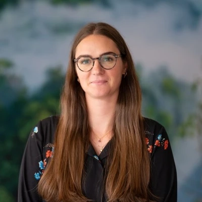 Woman with long brown hair in a dark floral shirt against a soft nature-themed backdrop