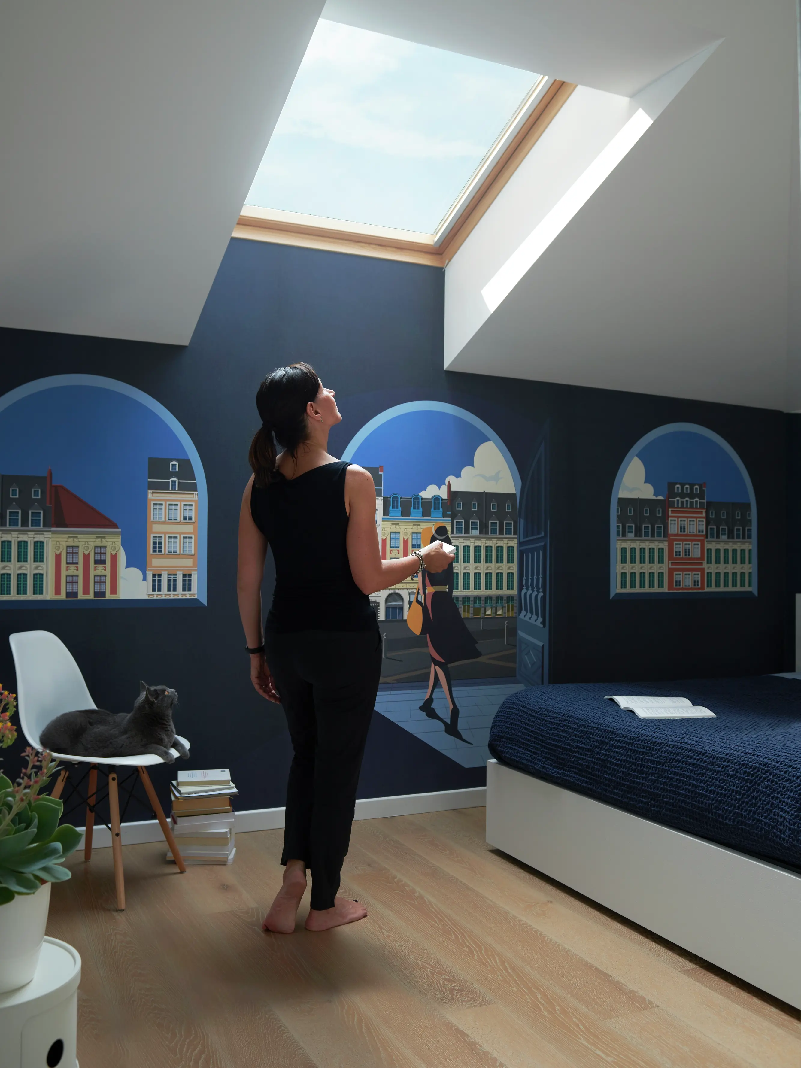 Camera da letto con murale di panorama urbano, finestra per tetti VELUX, gatto sulla sedia e copriletto blu navy.
