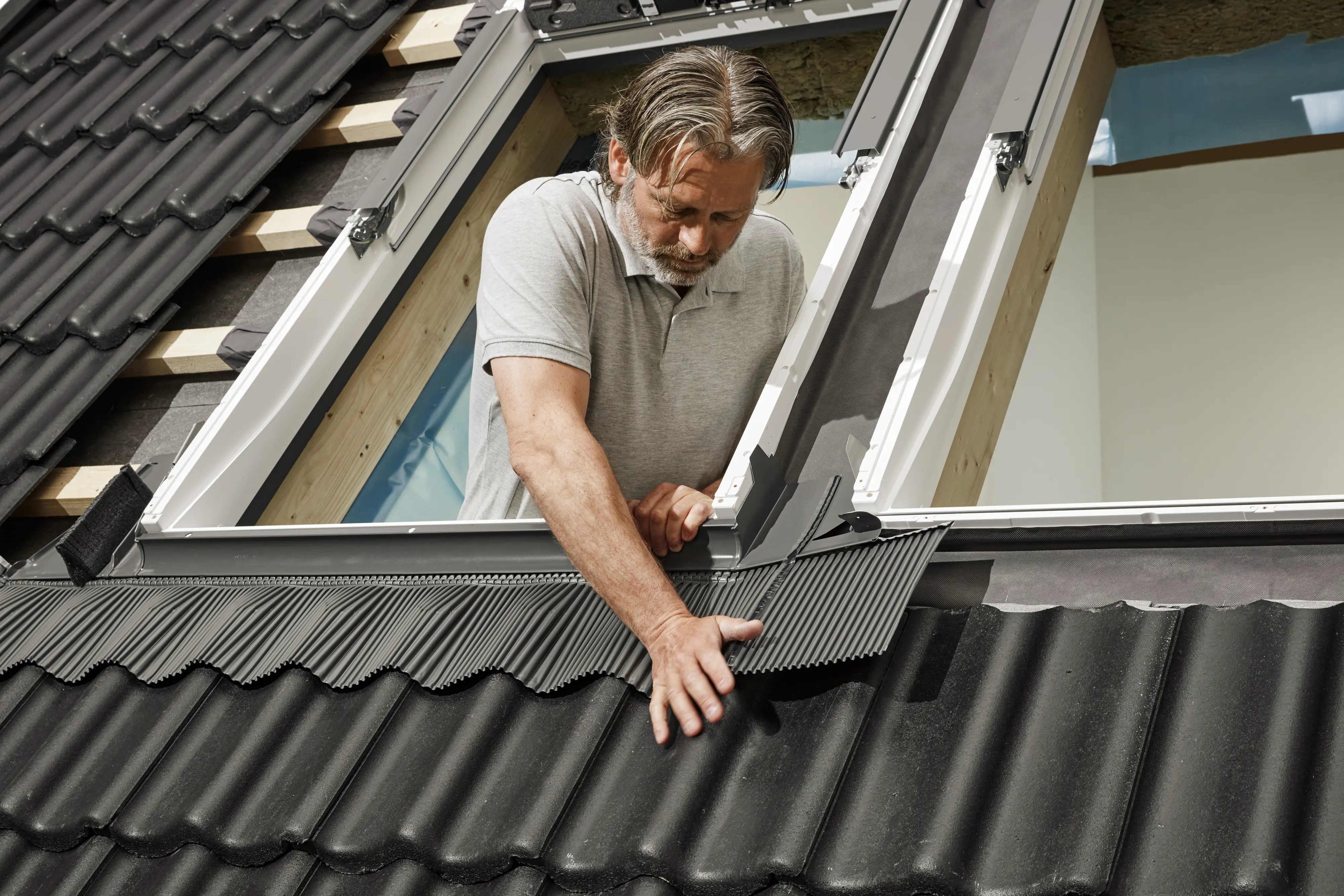Személy VELUX tetőablakot szerel fel egy fekete cserepekkel fedett, ferde tetőre.