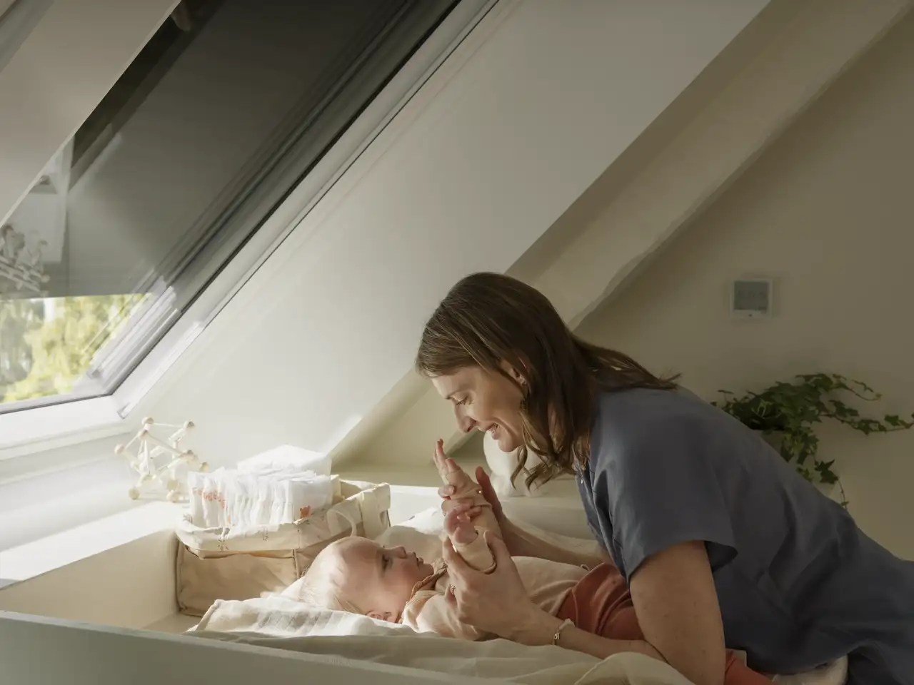 Nourrisson allongé sur une table à langer sous une fenêtre de toit VELUX équipée d’un volet roulant.