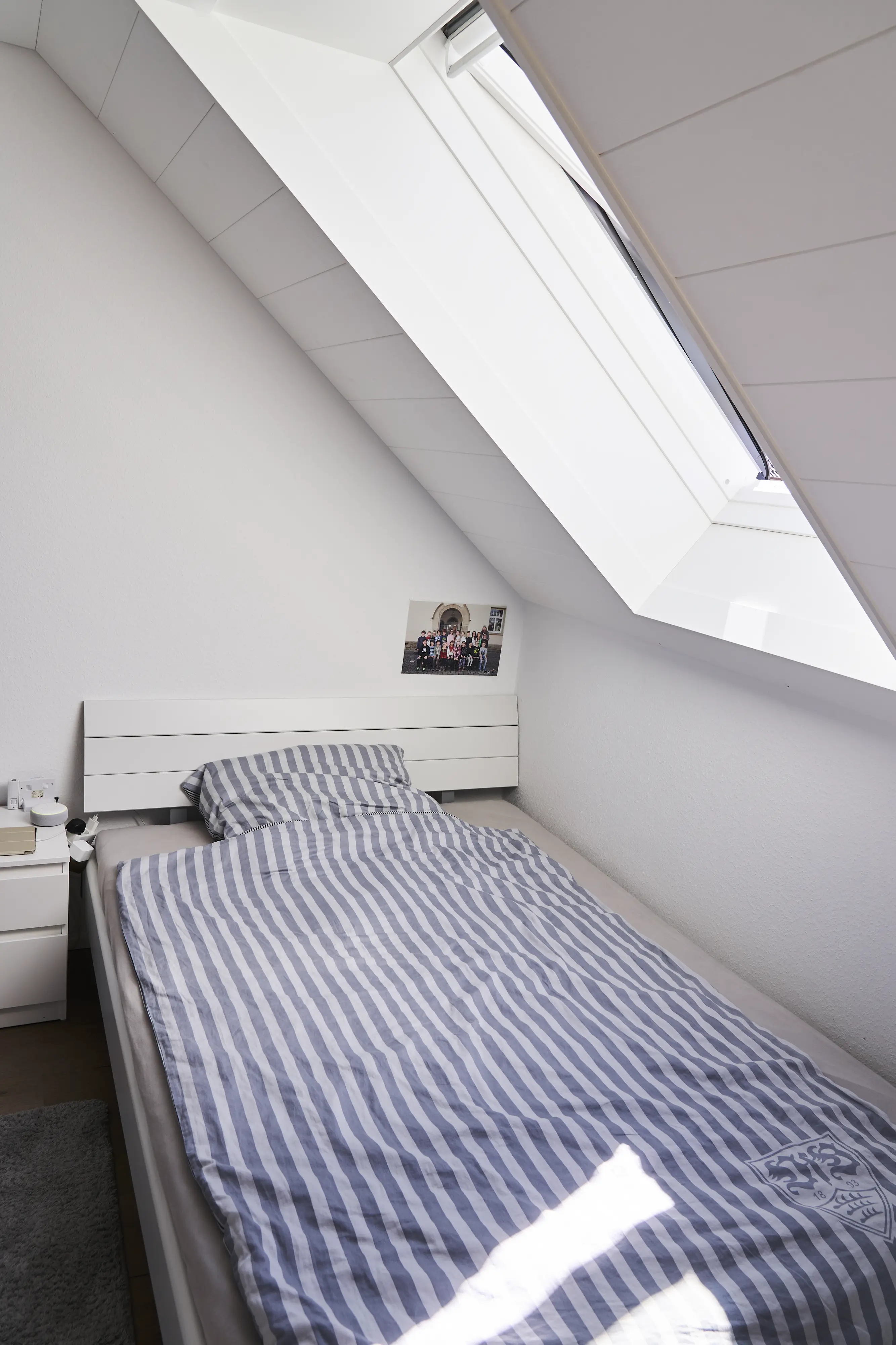 Bēniņu guļamistaba ar slīpiem griestiem un VELUX jumta logu, kā arī svītrainu gultasveļu.