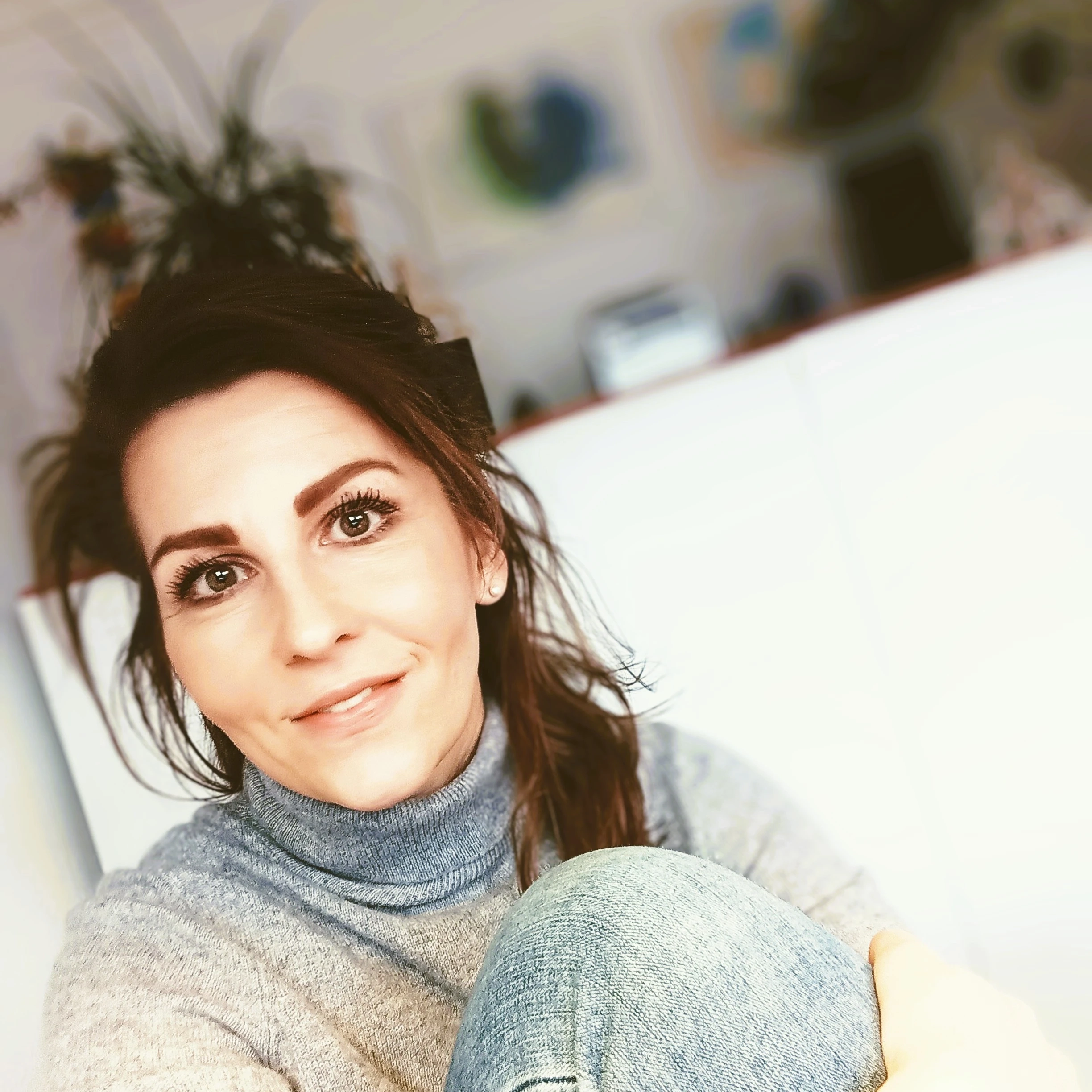 Person in grey sweater and jeans in a cozy indoor setting with blurred artwork.