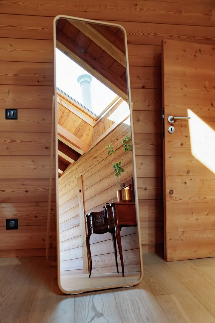 Drvena soba s prirodnim svjetlom koje se odbija u visokom ogledalu, stvarajući toplu i privlačnu atmosferu. | VELUX
