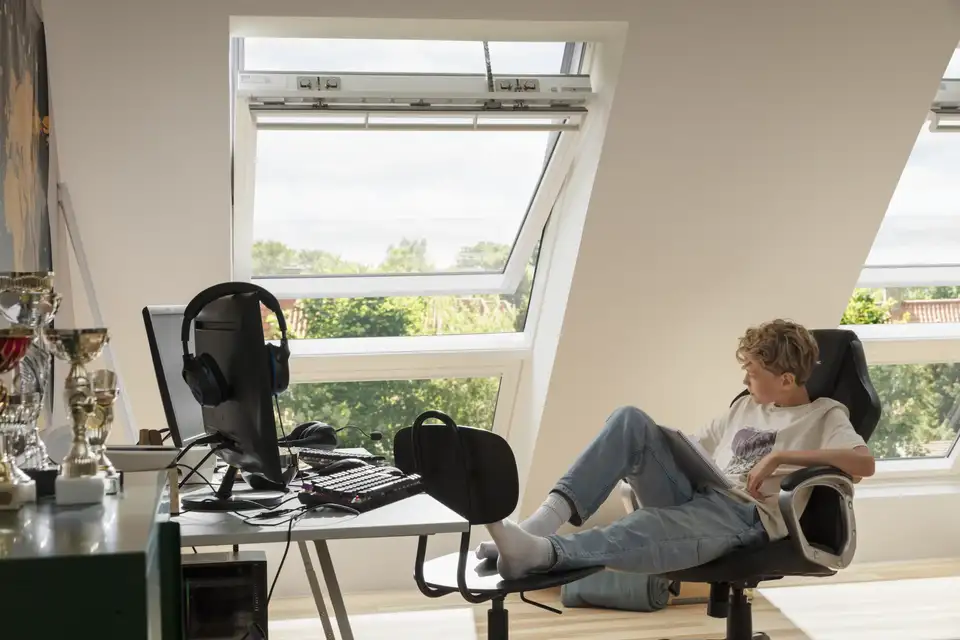 Garçon regardant vers l’extérieur à travers des fenêtres de toit VELUX, dans un bureau mansardé lumineux 
