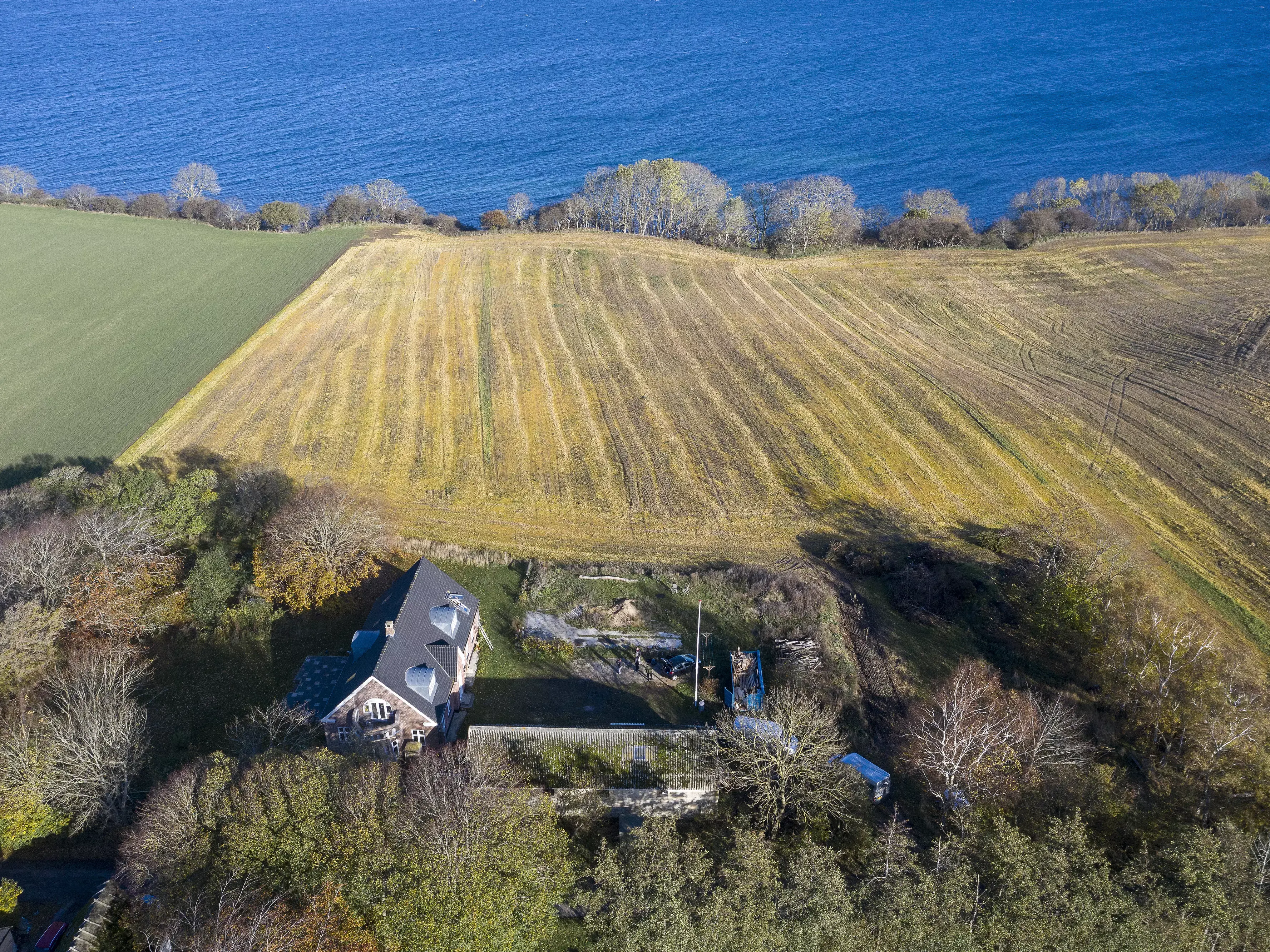 Vue aérienne d'une maison individuelle au bord de la mer, entourée d'arbres et de champs.