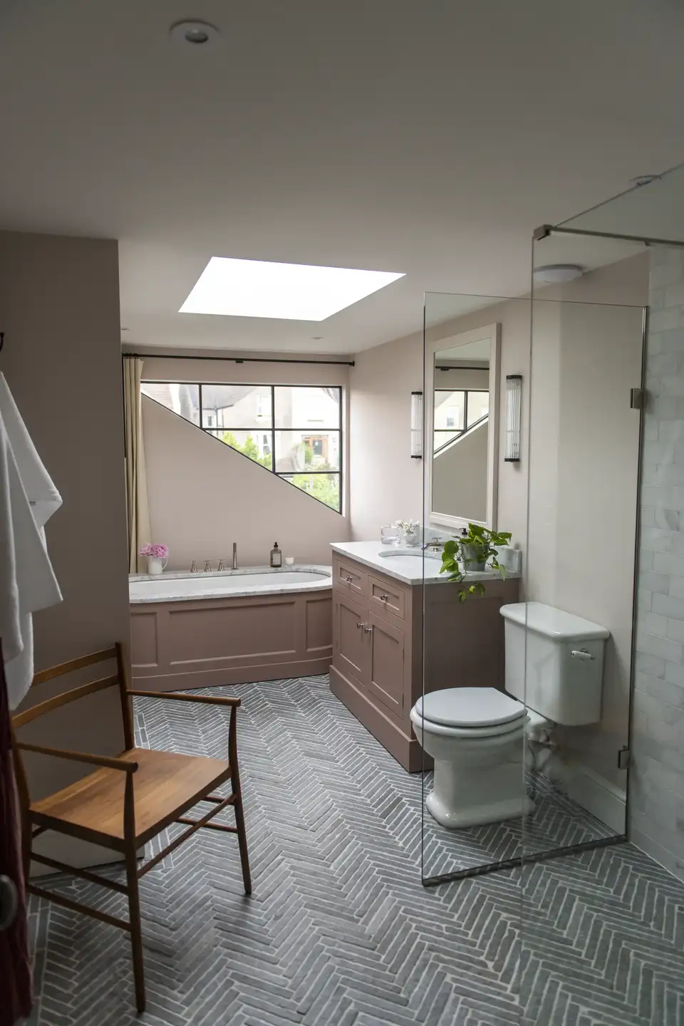 Une salle de bain avec des toilettes à droite, un lavabo avec meuble au centre, et une douche vitrée à gauche. Une chaise en bois est placée au centre sous un puits de lumière. Le sol est carrelé en motif chevrons et des plantes décorent le meuble.