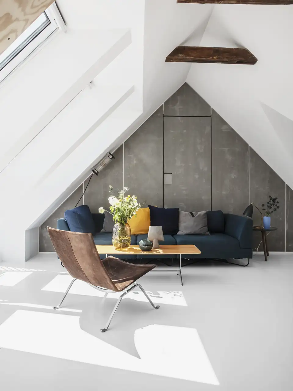 Grenier lumineux en style Bauhaus avec mur gris, canapé bleu et poutres apparentes.