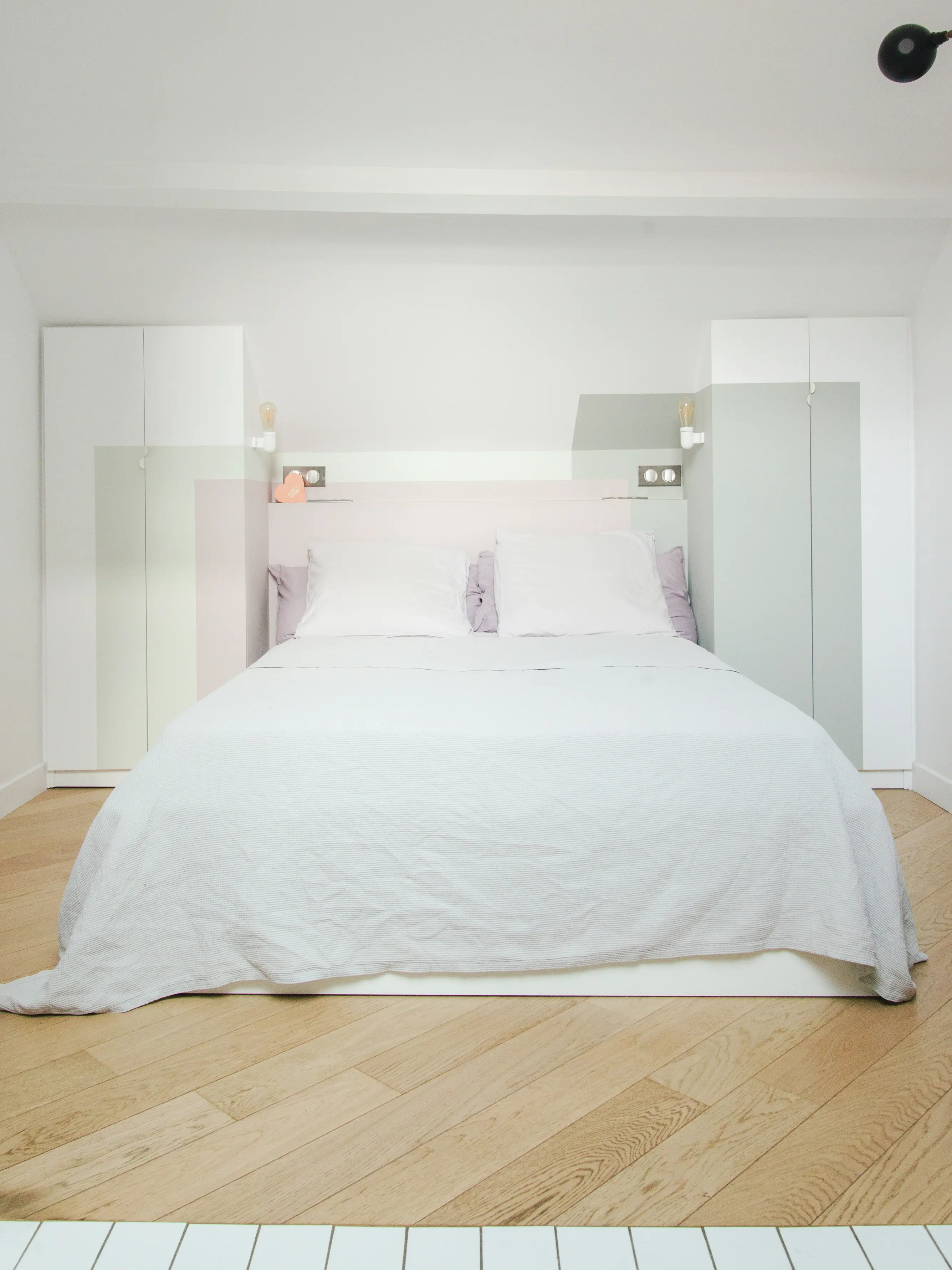 Camera da letto minimalista con biancheria da letto bianca, accenti color lavanda e pavimento a spina di pesce in legno.