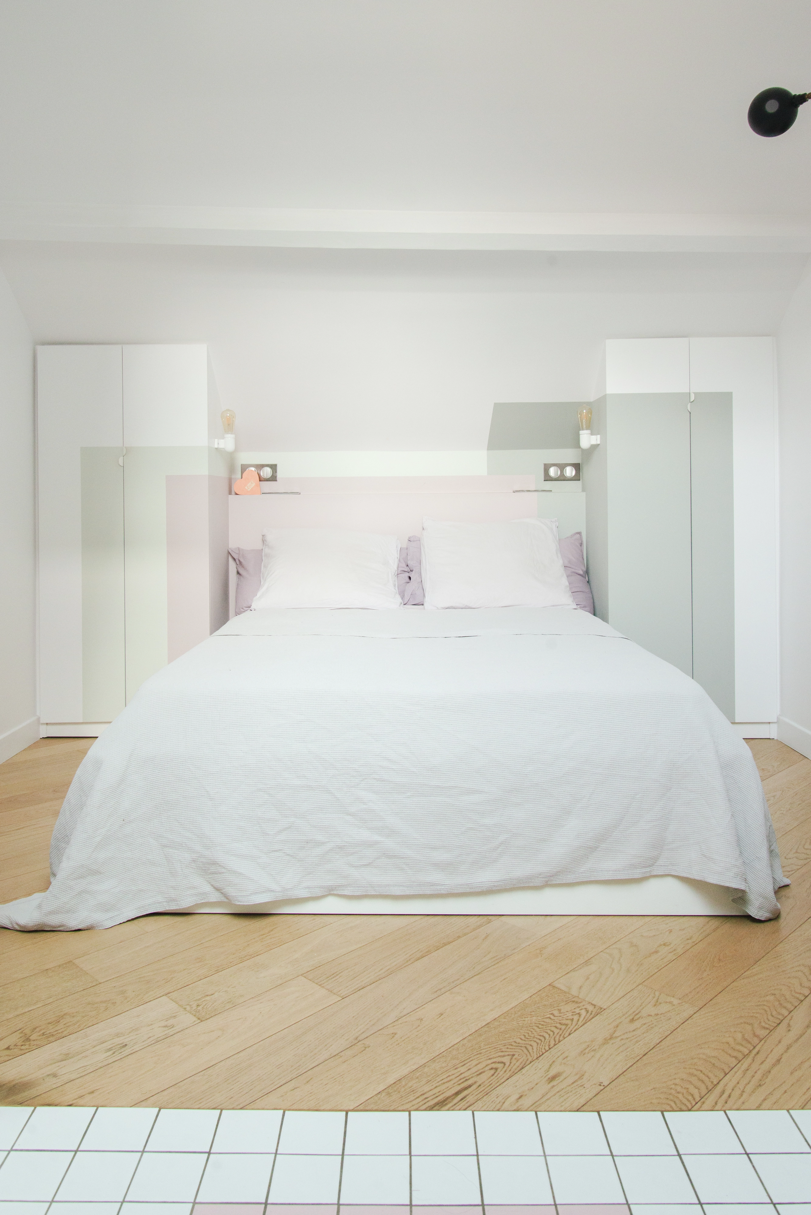Chambre minimaliste moderne avec décor blanc et rose pastel, sol en bois, et armoires blanches.