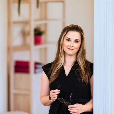 Woman leaning in a bright home office doorway, holding glasses and smiling softly