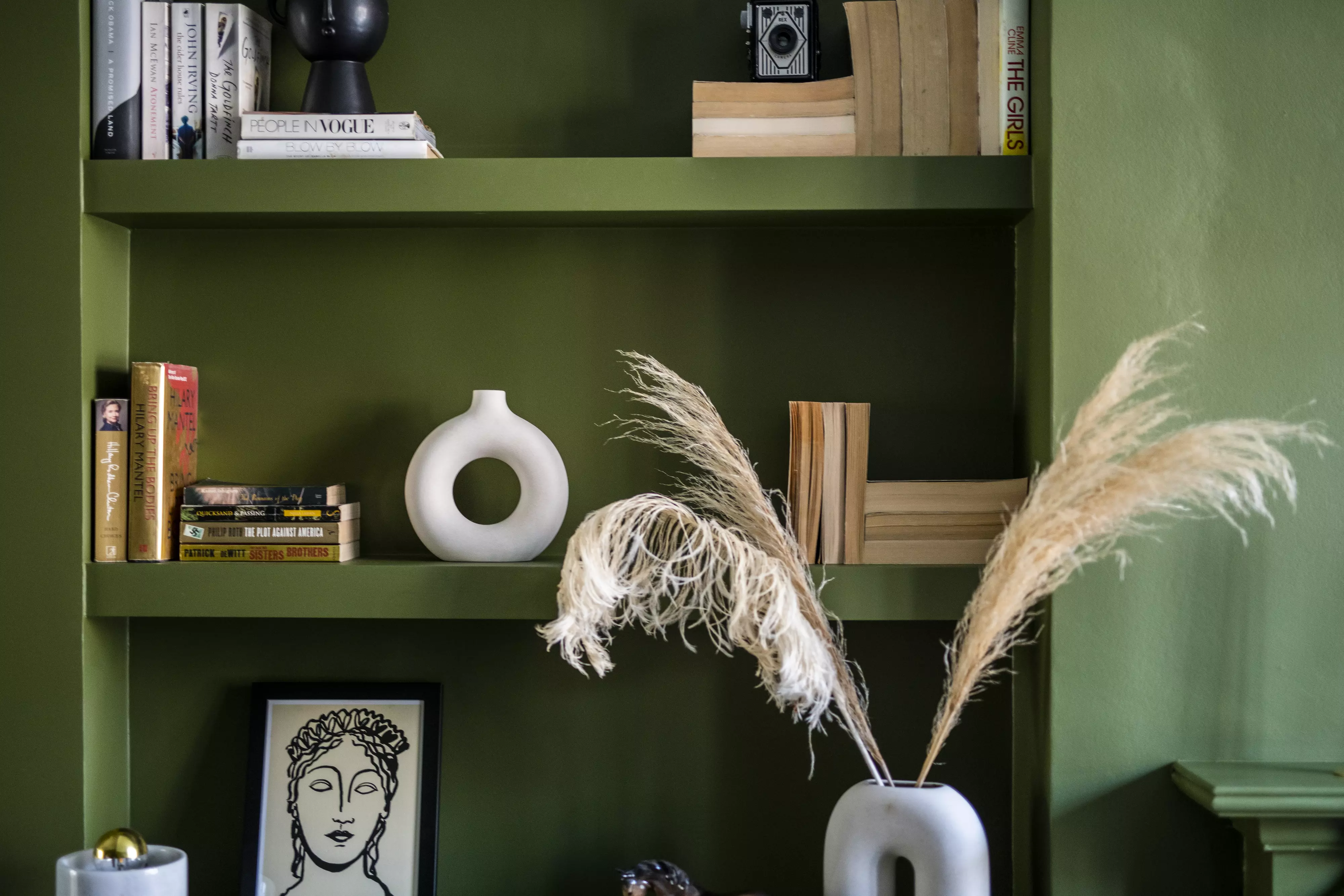 Home office shelf with books, decorative vases, and pampas grass against a green wall.