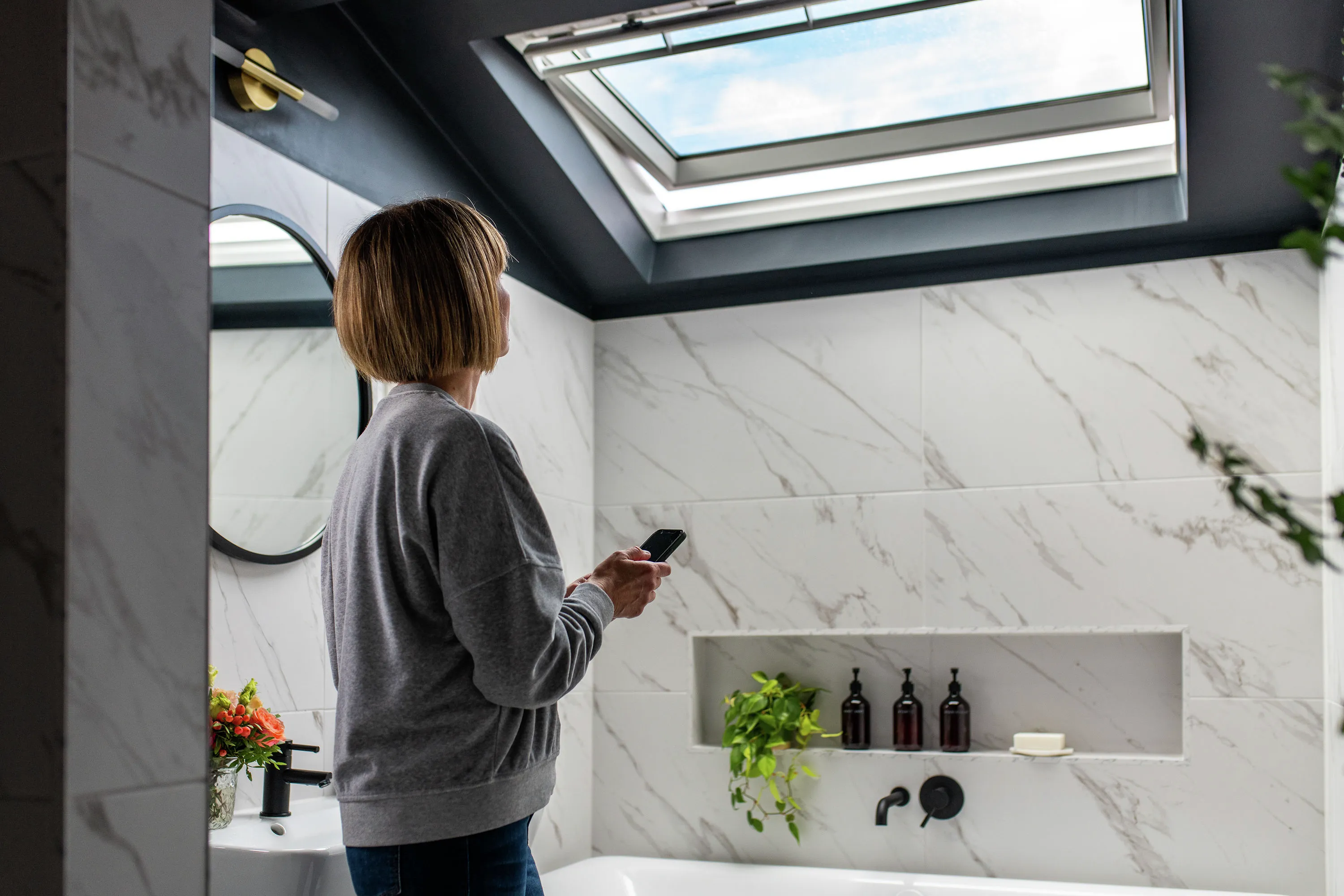 Egy pulóvert viselő nő okostelefonnal a kezében egy modern, világos fürdőszobában áll, és a nyitott, VELUX tetőablak felé néz, amivel természetes szellőztetés útján gondoskodik a gyors légcseréről.