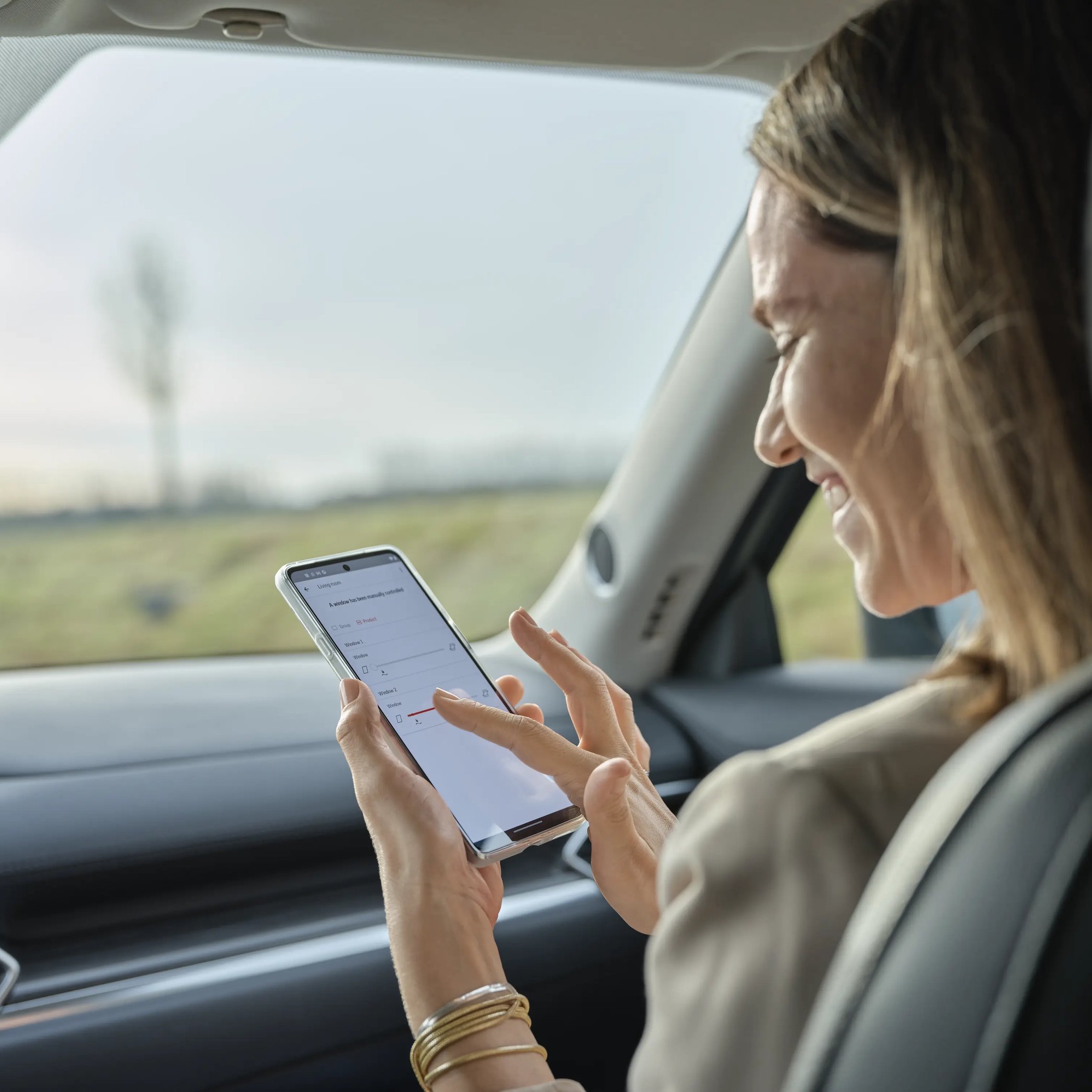Woman in car using smartphone, modern interior and rural landscape visible.