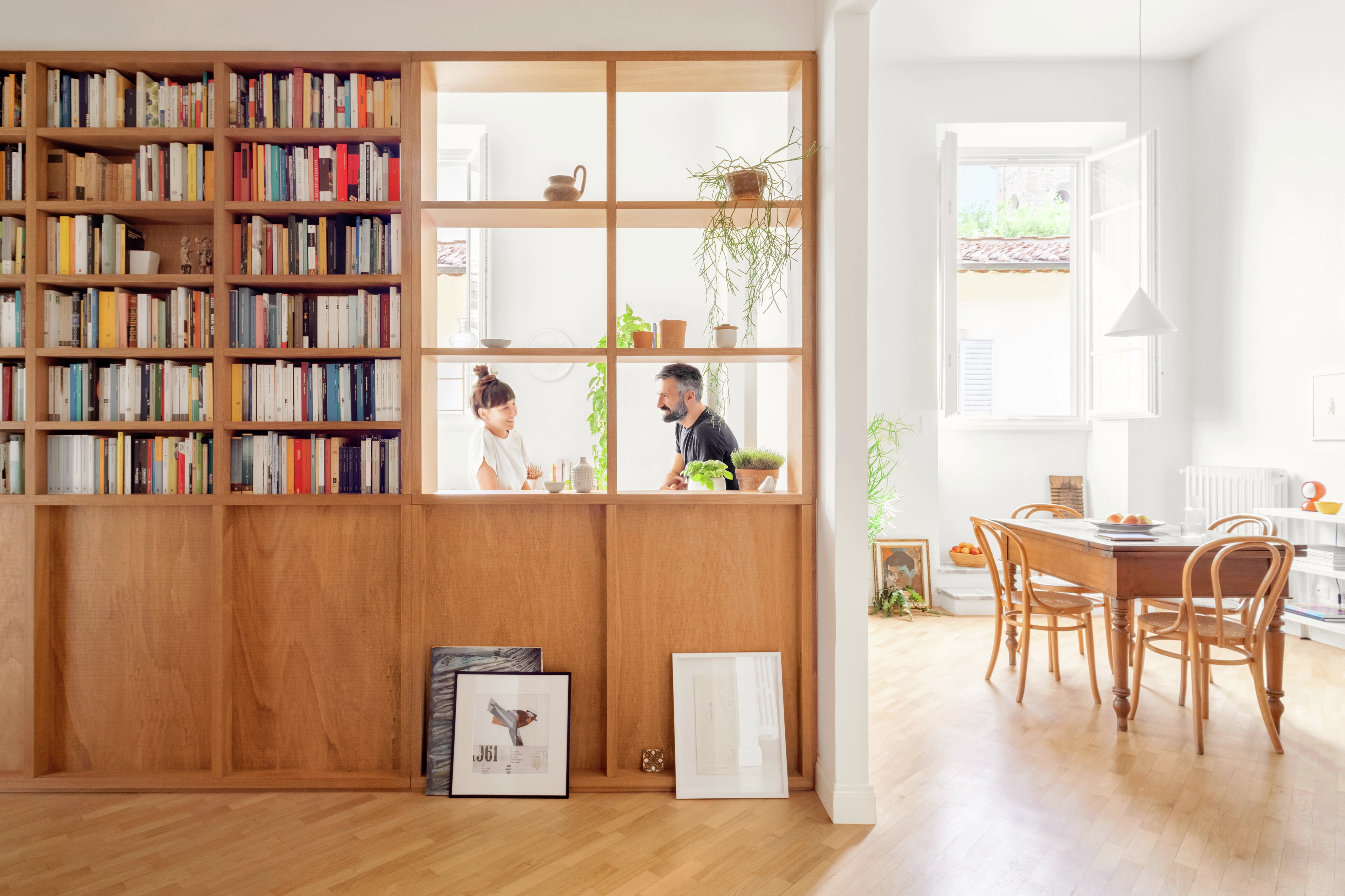 L’immagine mostra uno spazio interno luminoso e ben illuminato con grandi finestre. C’è una libreria di legno di notevoli dimensioni che copre la maggior parte del muro sul lato sinistro, riempita con vari libri e oggetti decorativi. Di fronte alla libreria, ci sono due immagini incorniciate appoggiate contro di essa sul pavimento. A destra, c’è un’area pranzo con un tavolo e delle sedie di legno. La stanza ha pavimenti in legno duro di colore chiaro e sembra essere ordinata, dando un’atmosfera accogliente e invitante.