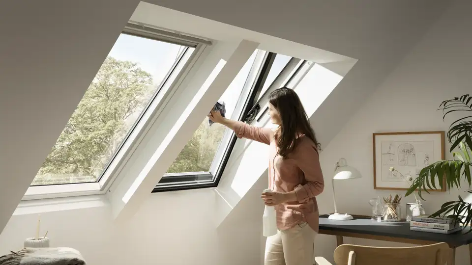 Person cleaning a VELUX roof window in a bright attic room.