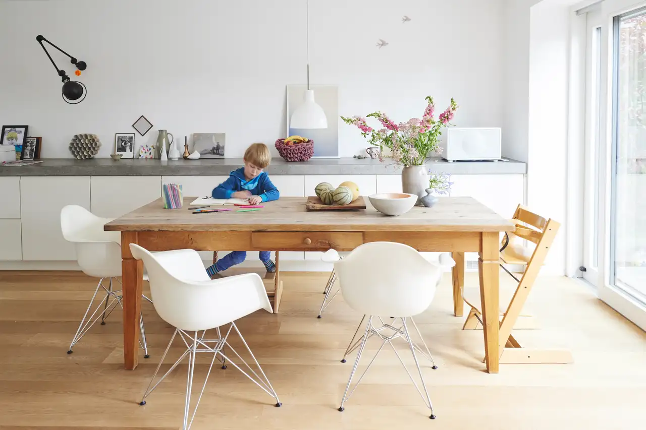 Cuisine lumineuse avec table en bois, chaises blanches et un enfant dessinant assis.