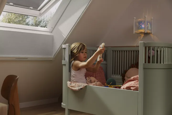 Kleines Mädchen sitzt in ihrem Kinderbett neben einem offenen Dachfenster mit Insektenschutzgitter | VELUX Magazin