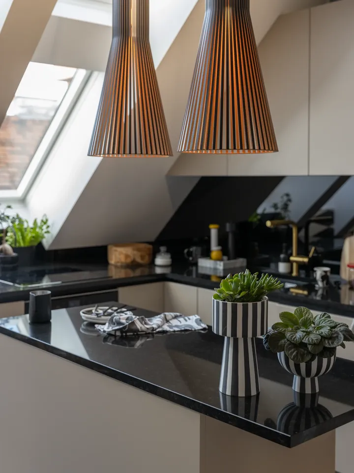 Offene Küche unter Dachschräge mit Hängeleuchten, cremefarbenen Fronten, dunkler Arbeitsplatte und Lichteinfall durch Dachfenster | VELUX Magazin