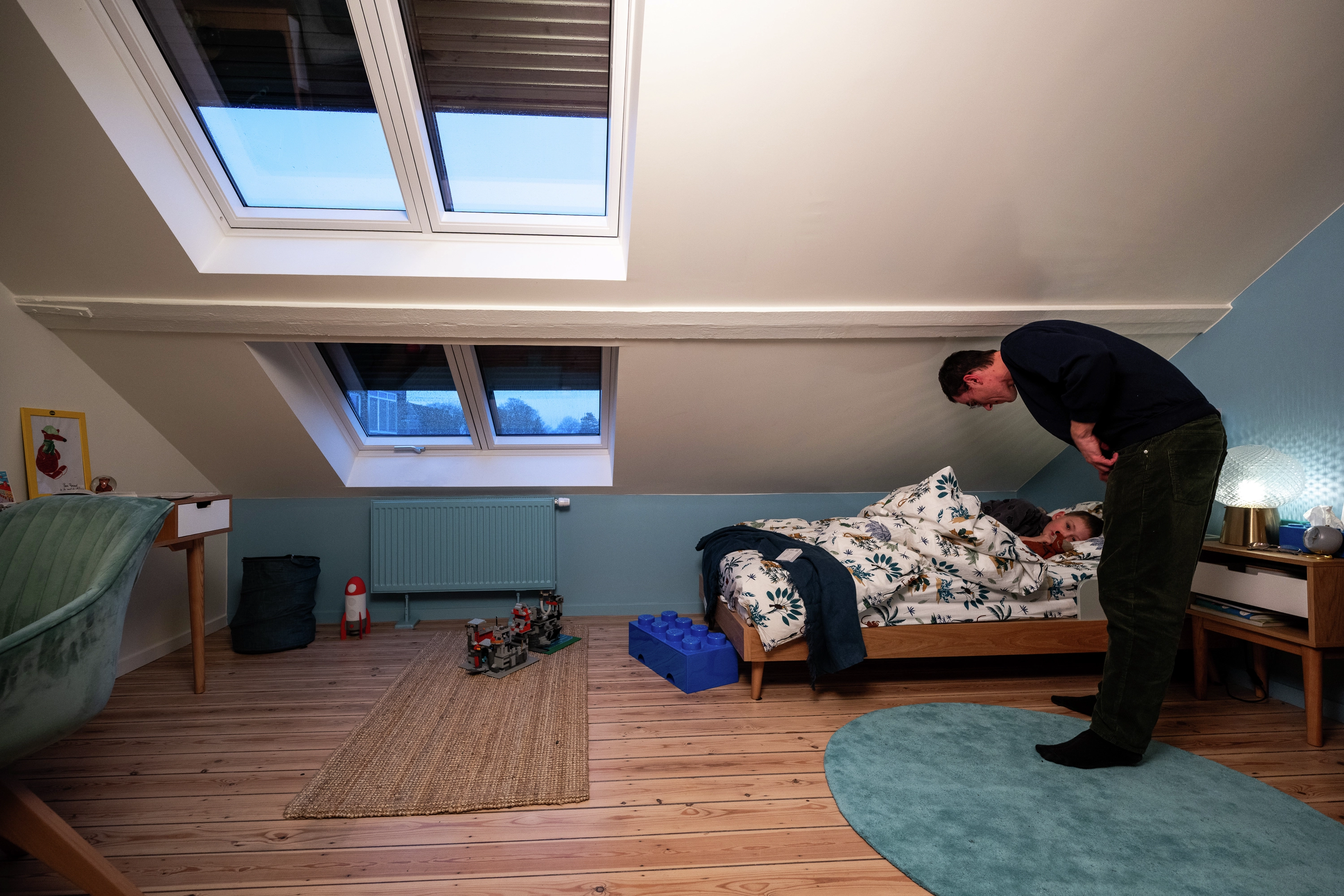 Vue de nuit : adulte bordant un enfant dans sa chambre, fenêtres de toit équipées de volets roulants.