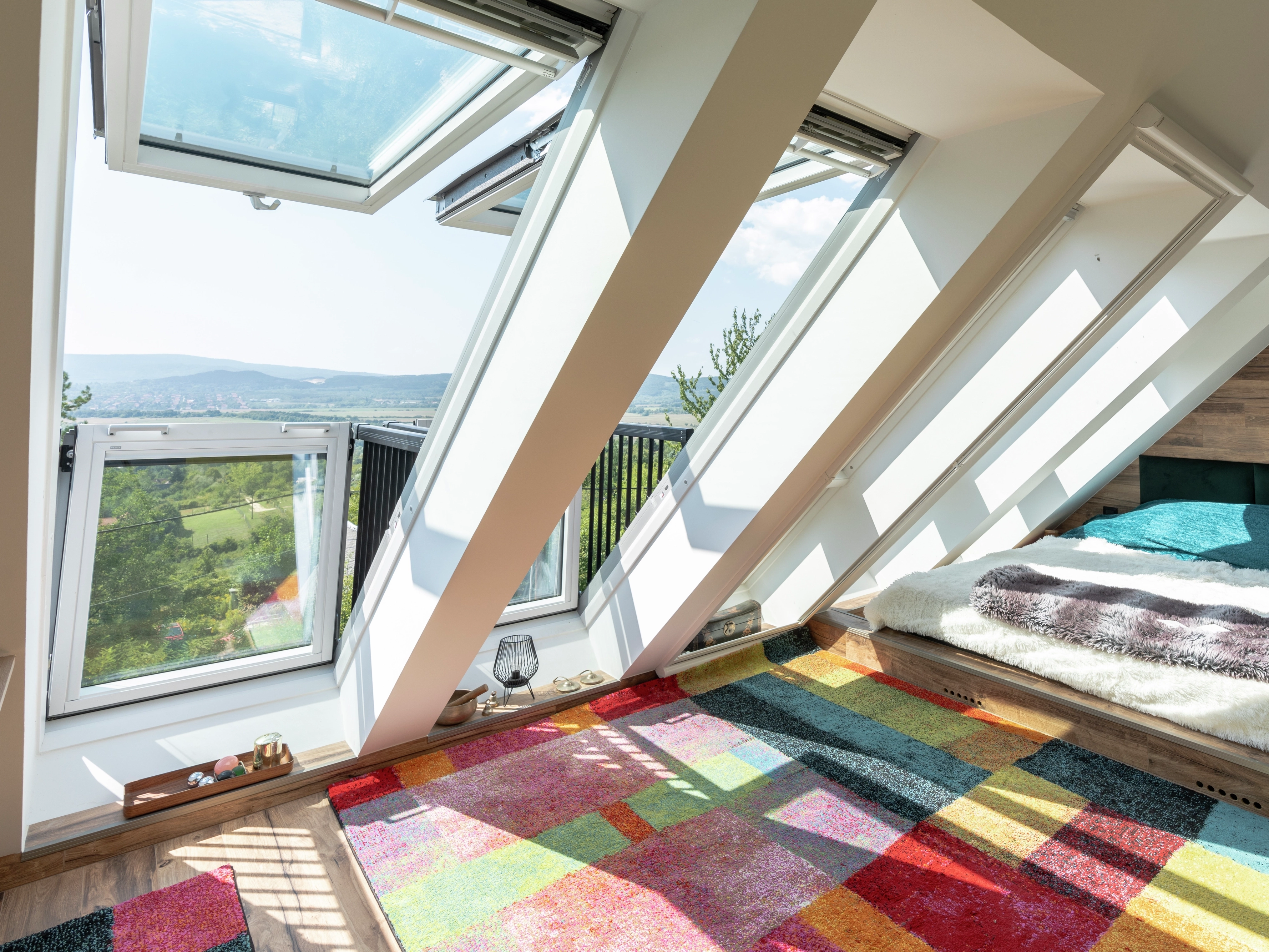 Attic bedroom with open VELUX roof windows, colorful rug, and scenic view.