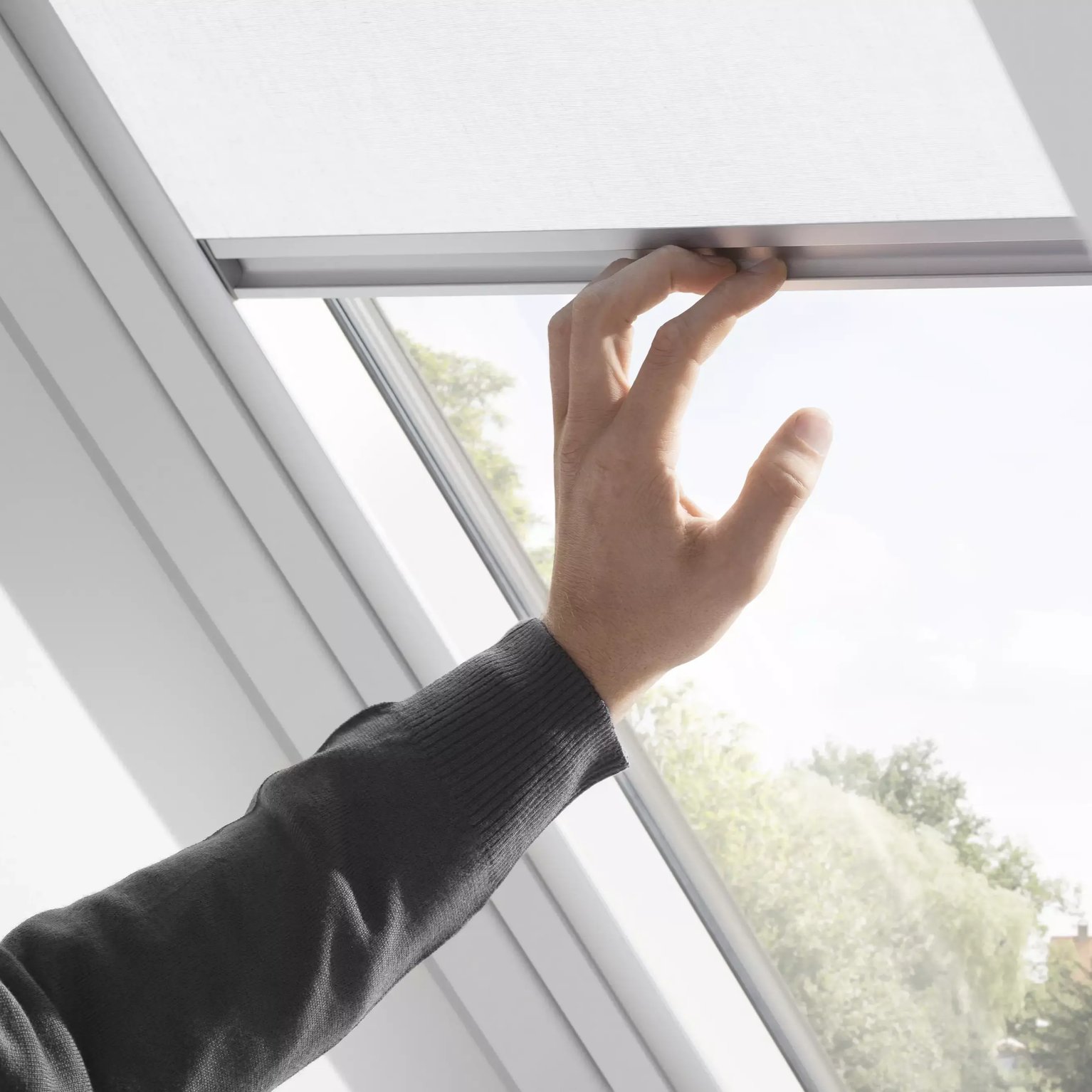 Hand adjusting blind on VELUX roof window, view of outdoors visible.