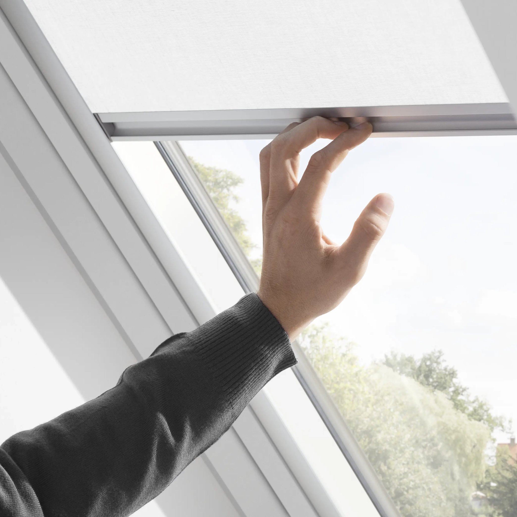 Hand adjusting blind on VELUX roof window, view of outdoors visible.