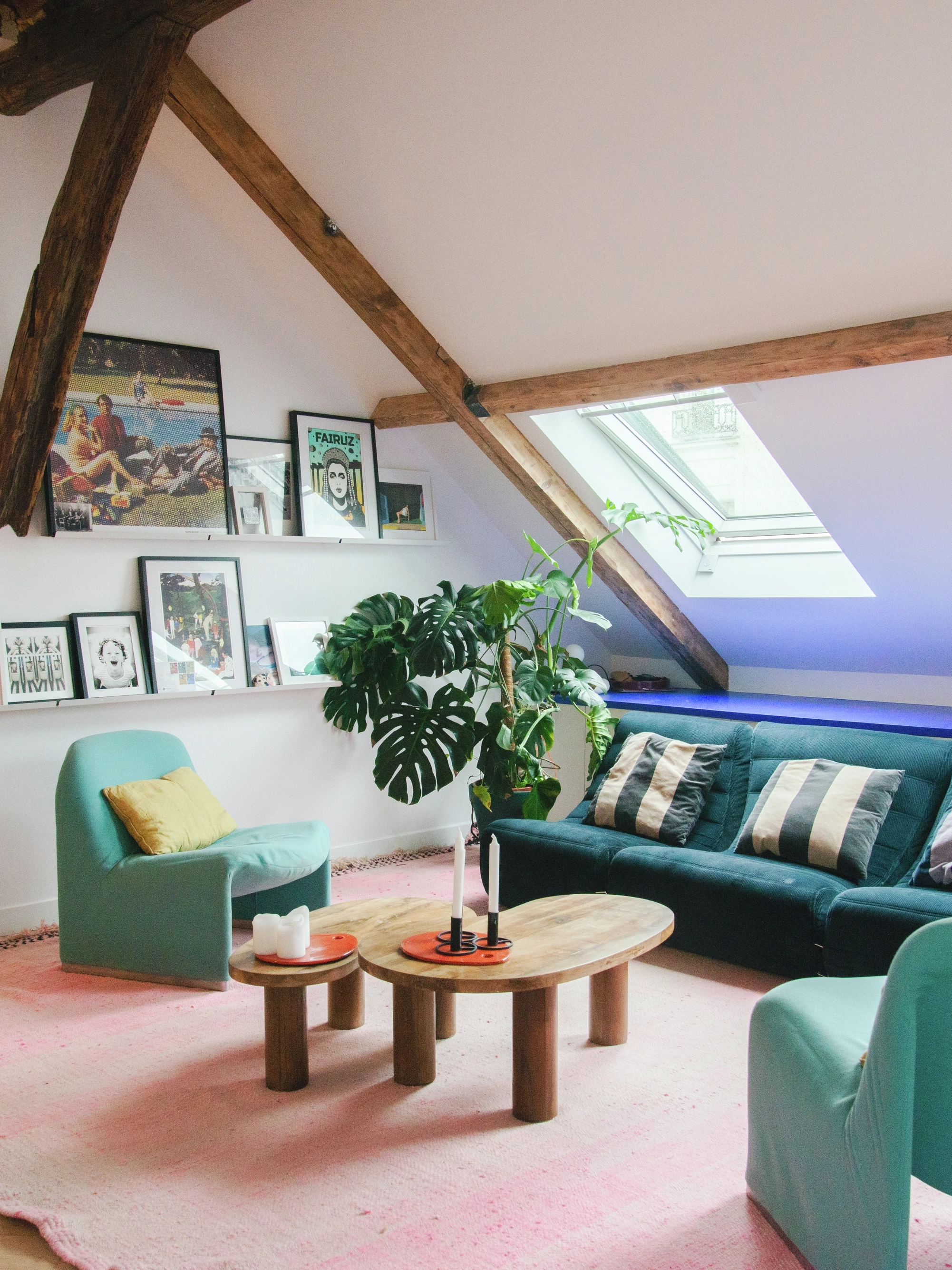 Une pièce mansardée avec deux fenêtres de toit Velux. Meublée d’un canapé bleu, deux fauteuils verts et une table basse. Décorée de photos encadrées et une plante en pot.